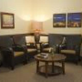 A cozy living room with comfortable chairs and a sofa, centered around a wooden coffee table. The space is softly lit by a lamp, with artwork adorning the walls.