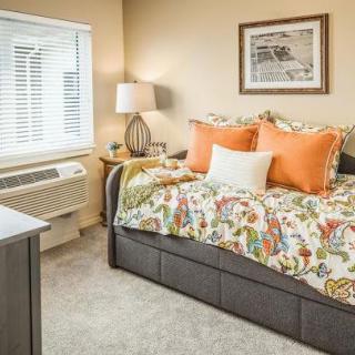 A cozy bedroom with colorful bedding and decorative pillows, featuring a bedside lamp and a window with blinds.