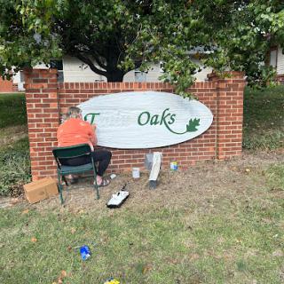 A person is painting a sign that says 'The Oaks' on a brick structure.