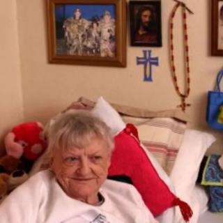 An elderly woman sitting comfortably in a bed surrounded by home decor and religious items.