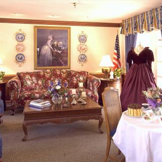 A vintage-themed living room featuring ornate furniture, a historic painting, and a dress on display, with an American flag in the corner.