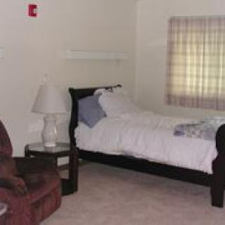 A cozy bedroom featuring a single bed, an armchair, and a bedside lamp, accompanied by a window with curtains.