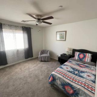 A cozy bedroom with a single bed featuring a patterned comforter, an armchair by the window, and a ceiling fan above.