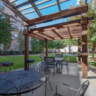 A sunny patio area with a wooden pergola, tables, and chairs surrounded by greenery and buildings.