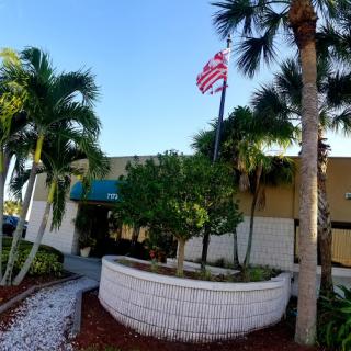 A sunny day showcasing a building surrounded by palm trees and greenery, with an American flag flying.