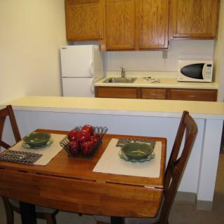 A compact kitchen and dining area featuring a wooden table set with dishes and a centerpiece, alongside basic appliances like a refrigerator and microwave.