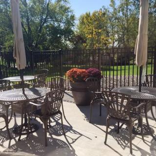 A sunlit patio with metal tables and chairs, surrounded by greenery.