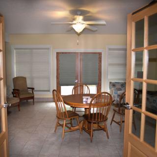 A cozy dining room with a wooden table and chairs, featuring large windows and a ceiling fan.