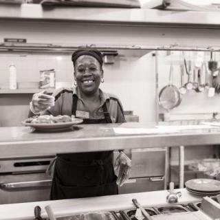 A chef happily serving food in a bustling restaurant kitchen.