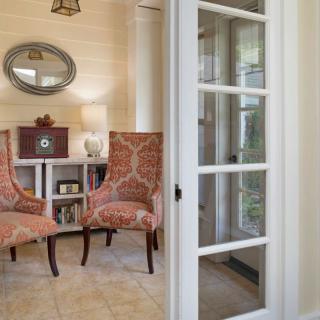 A cozy living room with two upholstered chairs and a round mirror, separated by a glass-paneled door.