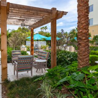 A peaceful garden patio featuring a wooden pergola and outdoor seating surrounded by lush tropical plants.