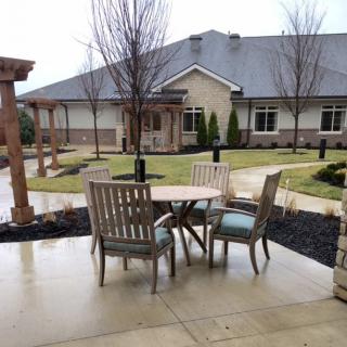 A patio with wooden chairs and a table overlooking a landscaped garden area in front of a modern building.