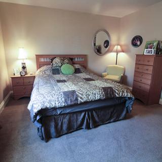 A cozy bedroom featuring a large bed with multiple pillows, two wooden dressers, lamps on either side of the bed, a round mirror on the wall, and carpeted flooring.