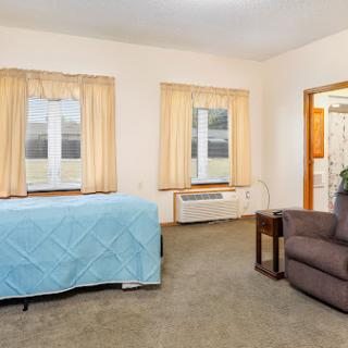 A cozy room featuring a bed with a blue coverlet, a comfortable chair, and large windows with light curtains.