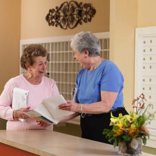 Two senior women engaged in a conversation at a reception desk, looking at paperwork together.