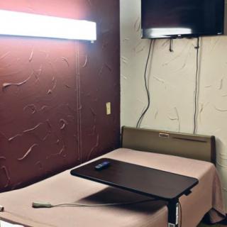 A simple room with a bed, wall-mounted television, and a small table under bright lighting.