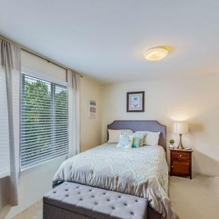 A cozy bedroom with a neatly made bed, large window with curtains, and a nightstand with a lamp.