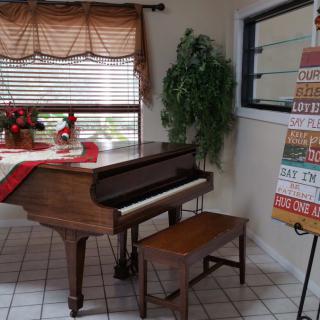 A cozy living room corner featuring a grand piano with a bench, decorated window, and an inspirational sign.