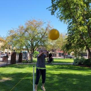 A person practicing balance on a slackline while reaching for a yellow ball in a sunny park.