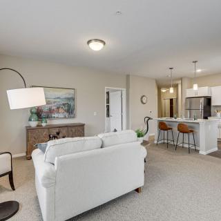 A modern open floor plan features a cozy living room with a white sofa, adjacent to a kitchen area with bar stools and pendant lighting.