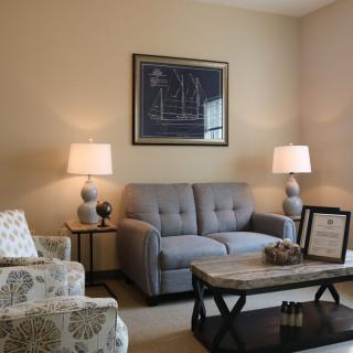 A cozy living room with a gray sofa, patterned armchair, wooden coffee table, and warm lighting from two lamps flanking the wall art.