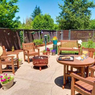 A sunny patio setting with wooden furniture, a fire pit, and lush green surroundings.