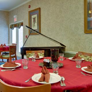 A dining room with a red tablecloth, set with glasses and napkins, featuring a piano in the corner.