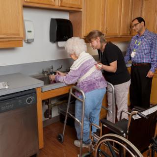 A caregiver assists an elderly woman using a walker in a kitchen setting, while another individual observes.