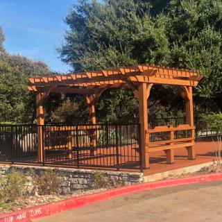 A wooden pergola with benches and a fence, set in a landscaped area with trees.