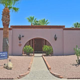 A charming desert home featuring a welcoming archway and surrounded by lush palm trees.
