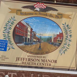 Welcome sign featuring a painted view of the town, sponsored by Jefferson Manor Health Center.