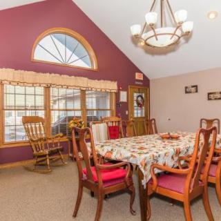 A cozy dining room with a large table set for a meal, adorned with a patterned tablecloth, surrounded by wooden chairs. The room features a vaulted ceiling with an elegant chandelier and large windows allowing natural light to fill the space.