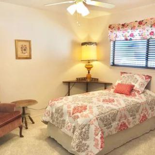A cozy bedroom featuring a single bed with floral bedding, an antique lamp, wall art, and a window with floral curtains.