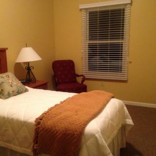 A cozy bedroom with a single bed, a lamp on a nightstand, an armchair, and a window with blinds.