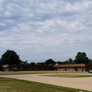 A view of a building under a cloudy sky, surrounded by grass and an empty road.