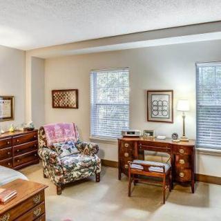 A cozy bedroom with classic furniture, featuring an armchair, a desk, and two large windows letting in natural light.