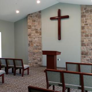 A small chapel interior featuring a cross on the wall, pulpit, and rows of pews.