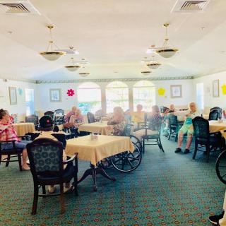 A bright dining room with elderly individuals seated at tables, some in wheelchairs.