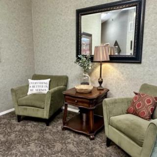 A cozy living room corner featuring green armchairs, a side table with a lamp, and a large mirror.