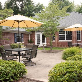 A cozy patio area with outdoor seating and yellow umbrellas, surrounded by lush greenery and a brick building.