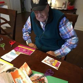An elderly man sitting at a table engaged in crafting activities with scissors and cards.