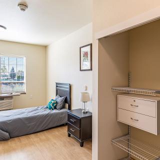 A minimalist bedroom with a single bed, nightstand, and an open closet, featuring wood flooring and natural light from the window.