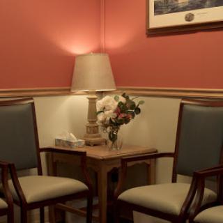 A cozy waiting area featuring two chairs, a small table with flowers, and a lamp, set against a warm pink wall.