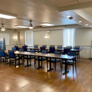 An empty conference room with arranged tables and chairs, featuring wooden flooring and overhead lighting.