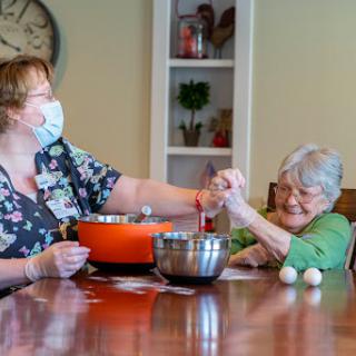 A caregiver and an elderly woman joyfully preparing a meal together at a dining table.