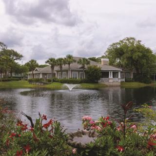 A beautiful house by a serene lake with a fountain, surrounded by lush greenery and colorful flowers under an overcast sky.