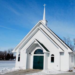 A small white church with a prominent steeple stands in a snowy landscape under a clear blue sky.