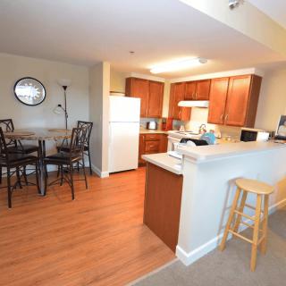 A modern apartment kitchen with dining area, featuring wooden cabinets and flooring.