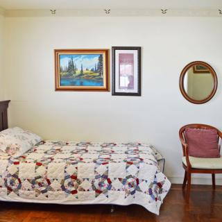 A simple bedroom with a single bed, decorated with a quilt, alongside paintings and a mirror on the wall, and a chair in the corner.