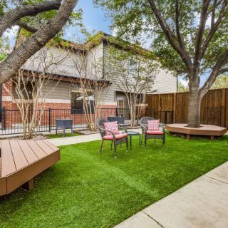 A cozy backyard with a lush green lawn, surrounded by trees and featuring several patio chairs and wooden benches.
